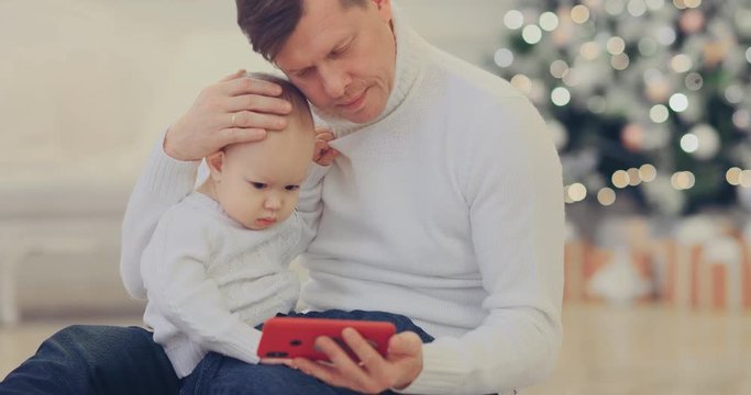 Kid sits with dad in his arms and watches a cartoon on the phone. Happy dad with little son sit together on the floor by the New Year tree. Portrait view.