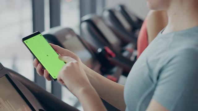 Athletic Woman Does Gestures while Using Green Screen Smartphone and Running on the Treadmill in a Gym. Fit Female Posts on Social Media, Takes Pictures, Watches Videos, Exercises in Fitness Club
