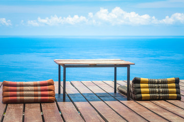 Fototapeta na wymiar Table and Thai Triangle Pillows on Wooden Terrace with Tropical Beach, Blue Sky and Clouds in Background