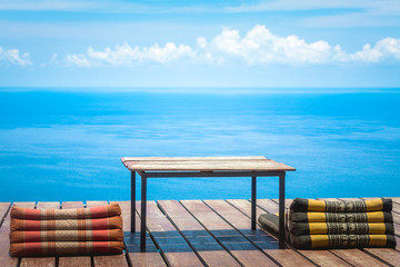 Table and Thai Triangle Pillows on Wooden Terrace with Tropical Beach, Blue Sky and Clouds in Background