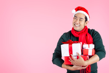 Happy Asian man wearing green sweater and Santa hat holding gift boxes on pink background and copy space.