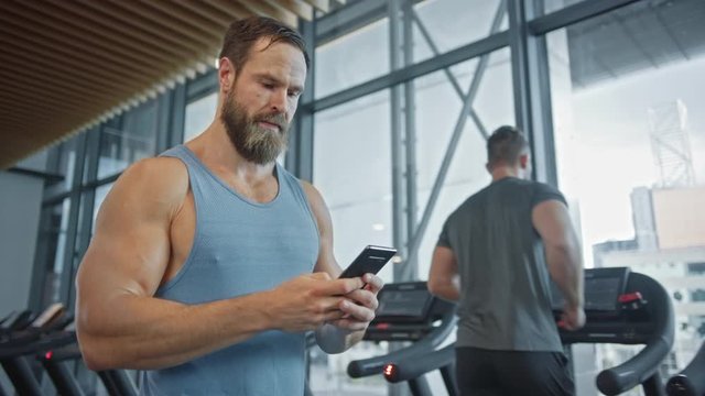 Muscular Heavyweight Champion Walks Through Gym, Uses Smartphone for Social Media and Conducting Business Affairs. In the Background Sports People Running on Treadmills