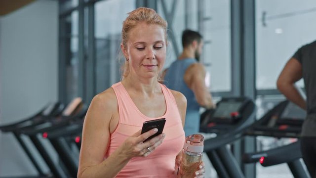 Beautiful Athletic Woman Uses Smartphone Walks Through Fitness Club Gym. Using Social Media, Posting Pictures, Communicating, Checking Email. In the Background Sports People Running on Treadmills