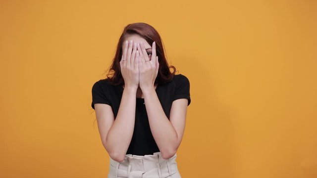 Attractive young woman in black shirt covering face with hands, looking through fingers isolated on orange background in studio. People sincere emotions, health concept.