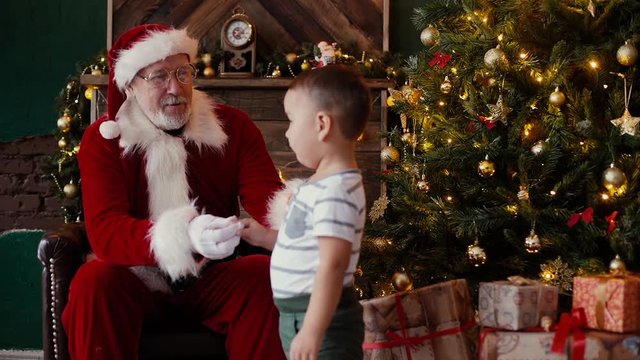 Little boy comes to the smiling Santa Claus, who sits next to the Christmas tree in room with festive interior. Shooting in slow motion.
