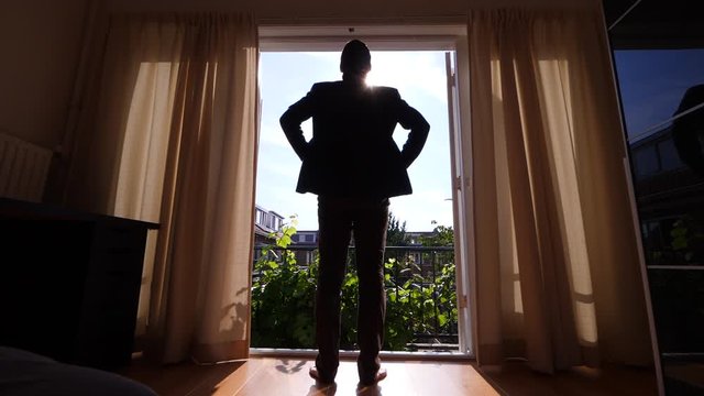 Man wearing causal suit stand against open doors, looking out to inner yard of his house. Guy hold arms on hips, stay still. Green leaves on small balcony, curtains slightly move by wind