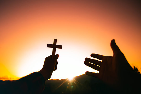 Human hand holding christian cross and praying with a sunset sky background. Christian silhouette concept.
