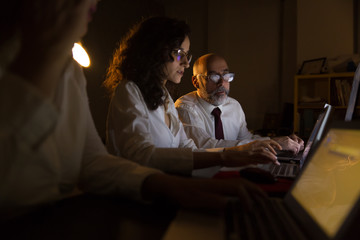 Businessman and businesswomen working with laptops. Side view of professional male and female business colleagues using laptop computers in dark office. Working late concept