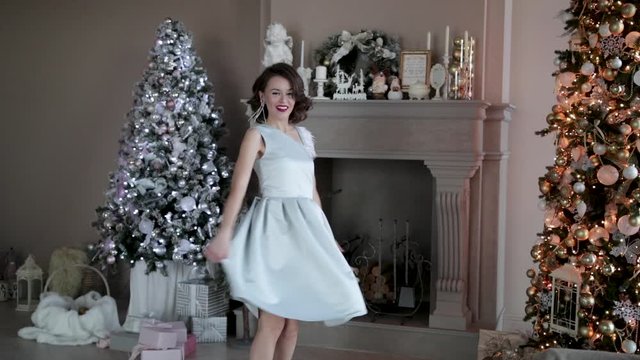 Beautiful sexy girl in a beautiful dress next to the Christmas tree on the background of the fireplace posing and walking at home. Woman spinning and dancing near the Christmas tree, gifts and good