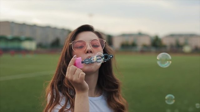 Beautiful young woman in sunglasses blowing soap bubbles outdoors and spinning around. Close view. Slow motion