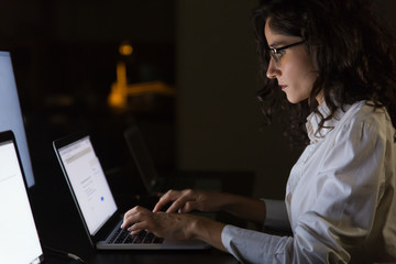 Serious businesswoman using laptop in dark office. Side view of professional young businesswoman...