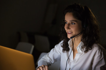 Smiling woman with headset in dark office. Cheerful female call center worker working with laptop...