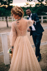 Young bride stands with her back to the camera and gives a hand to her groom, sunset at the ranch