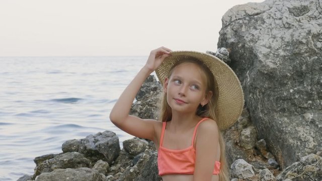 Cute teenager girl in bikini trying straw hat on stone in sea water. Portrait beautiful girl in summer hat on sea water landscape on rocky beach