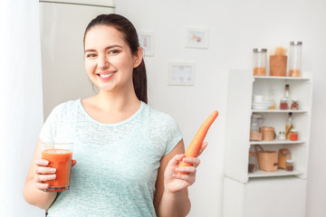 Fototapeta premium Body Care. Chubby girl standing in kitchen with glass of juice and carrot smiling happy close-up
