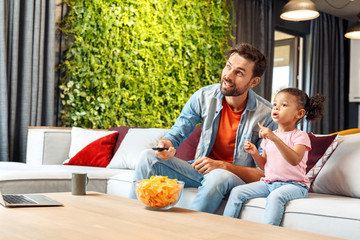 Mixed Race Family. Father and daughter sitting on sofa at home watching cartoon on tv together...