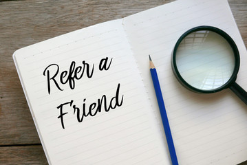 Top view of magnifying glass,pencil,and notebook written with Refer A Friend on wooden background.