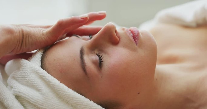 Close up of a beautiful young smiling woman with a towel on her head is receiving a facial massage and spa treatment for perfect skin in a luxury wellness center.