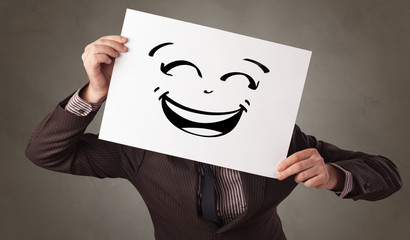 Casual person holding a paper in front of his face with drawn emoticon face