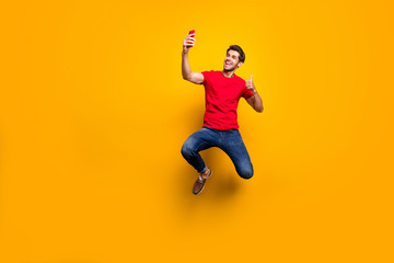 Full size photo of cheerful brown hair guy enjoy spring time holidays jump make selfie show thumb-up gesture recommend journey wear red t-shirt denim jeans isolated yellow color background