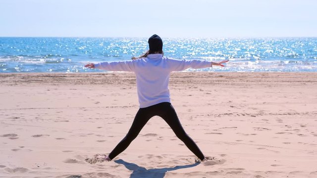 Barefooted woman beginner does triangle yoga pose on sand sea beach, back view. She is wearing hoodie, hat and leggings.