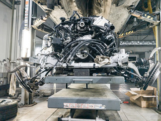 A used car raised on a lift for repair and under it a detached engine and gearbox suspended on a...