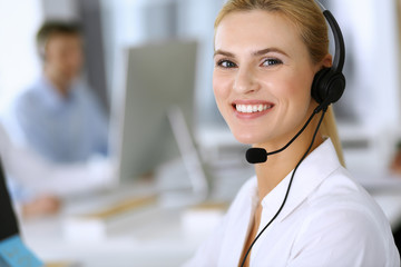 Blonde business woman using headset for communication and consulting people at customer service office. Call center. Group of operators at work at the background