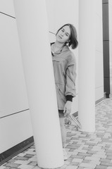 Black and white shot of A short haired woman in coral windbreaker and colorful socks hiding behind a column near grey wall