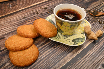 A Cup of black tea, tea leaves, pieces of brown sugar, oatmeal cookies on a wooden background. Close up.