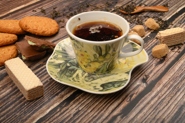 A Cup of black tea, tea leaves, pieces of brown sugar, oatmeal cookies, waffles, pieces of chocolate on a wooden background. Close up.