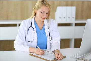 Doctor woman at work while sitting at the desk in hospital or clinic. Blonde cheerful physician filling up medical records form. Data and best service in medicine and healthcare