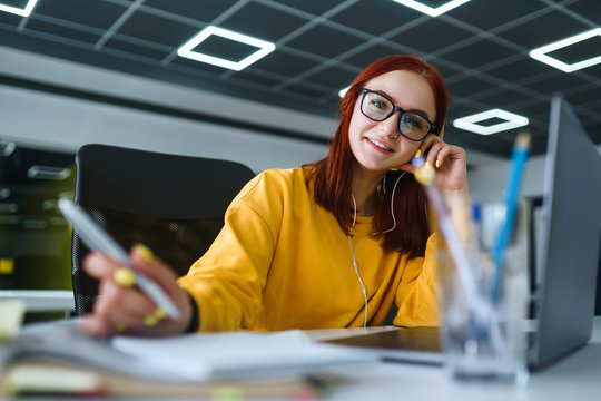 Young girl works at a computer in the office. Beautiful freelancer working from a laptop computer. Teenager student at the computer listens music to earphones and prepares for exams. Business woman.