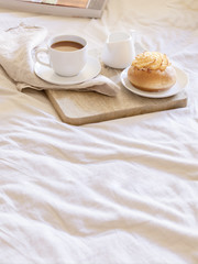 Good morning scenery background. Breakfast In Bed,  cup of coffee with sweet bun on wooden tray  and book on beige white sheets.