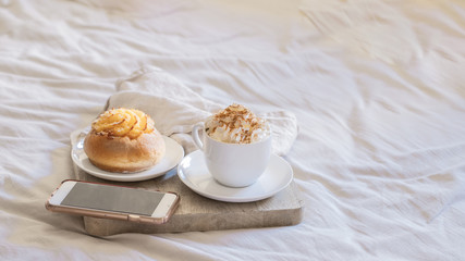 Morning Breakfast In Bed ,  cup of coffee  with foam milk and sweet bun,smartphone on wooden tray on beige white sheets. Good morning scenery background. Scandinavian style.