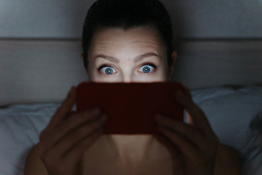 Girl addictively staring at her smartphone late at night in bed. Surprised woman face. Smartphone overuse. Technology and Imternet dependence concept.