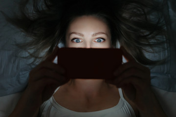 Young female addictively staring at her smartphone late at night in bed. Focused on eyes. Phone dependence concept. Horizontal shot. - obrazy, fototapety, plakaty