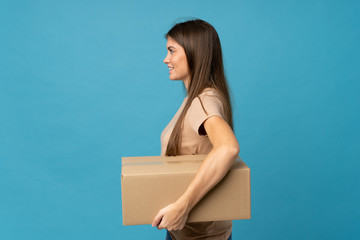 Young woman over isolated blue background holding a box to move it to another site in lateral position