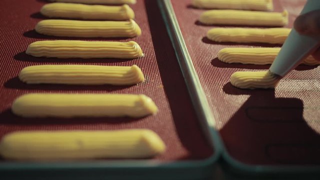 Making traditional French dessert, profiteroles, squeezing from pipe on tray. Uncooked eclairs lay on pan in lines, looking tasty.
