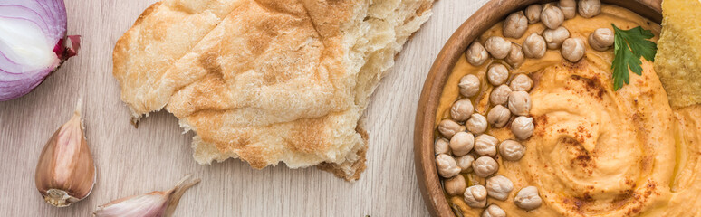 top view of delicious hummus with chickpeas in bowl near fresh baked pita and spices on beige wooden table, panoramic shot