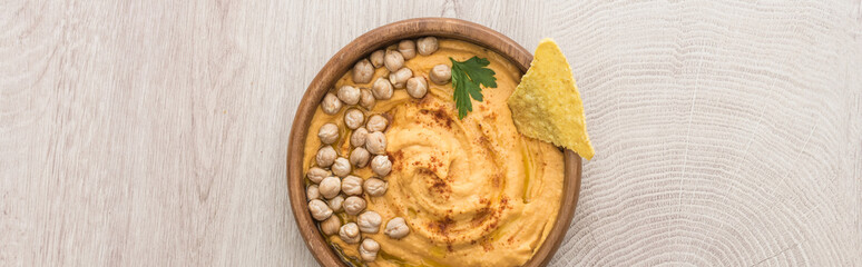 top view of delicious hummus with chickpeas and nacho in bowl on beige wooden table, panoramic shot