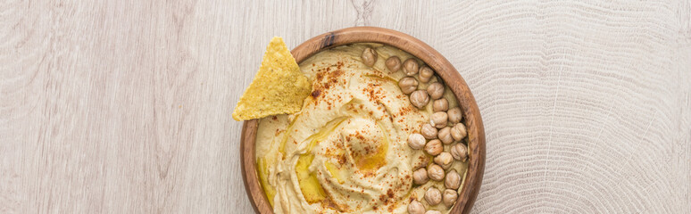 top view of delicious hummus with chickpeas and nacho in bowl on beige wooden table, panoramic shot