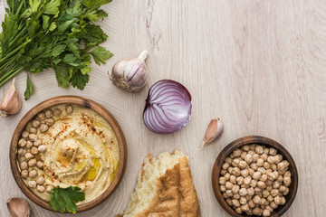 top view of delicious hummus with chickpeas in bowl near fresh baked pita, garlic, parsley and red onion on beige wooden table