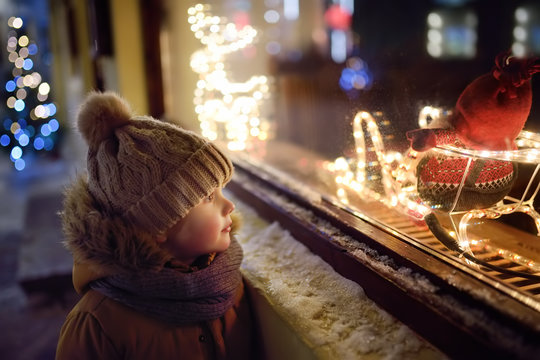 Little boy admires Christmas decorations in showcase of shop on winter evening. Tourist looking on Xmas toys and accessories on traditional Christmas market in Tallinn.