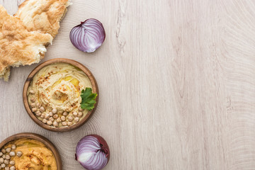 top view of delicious hummus with chickpeas in bowl near fresh baked pita, red onion on beige wooden table