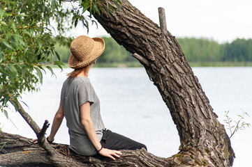 Red haired cute girl in summer outfit seats on the tree trunk near the lake  and looking on the...