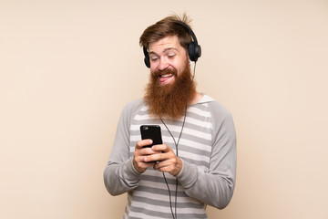 Fototapeta premium Redhead man with long beard over isolated background using the mobile with headphones and singing