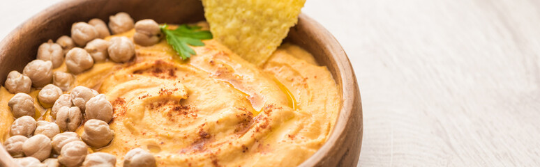 close up view of delicious hummus with chickpeas and nacho in bowl on beige wooden table, panoramic shot