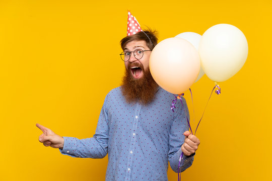 Redhead man with long beard holding balloons over isolated yellow background surprised and pointing side