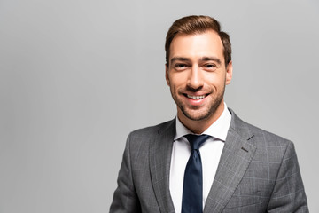 handsome and smiling businessman in suit looking at camera isolated on grey