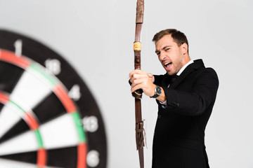 selective focus of angry businessman in suit holding bow and shooting at target isolated on grey